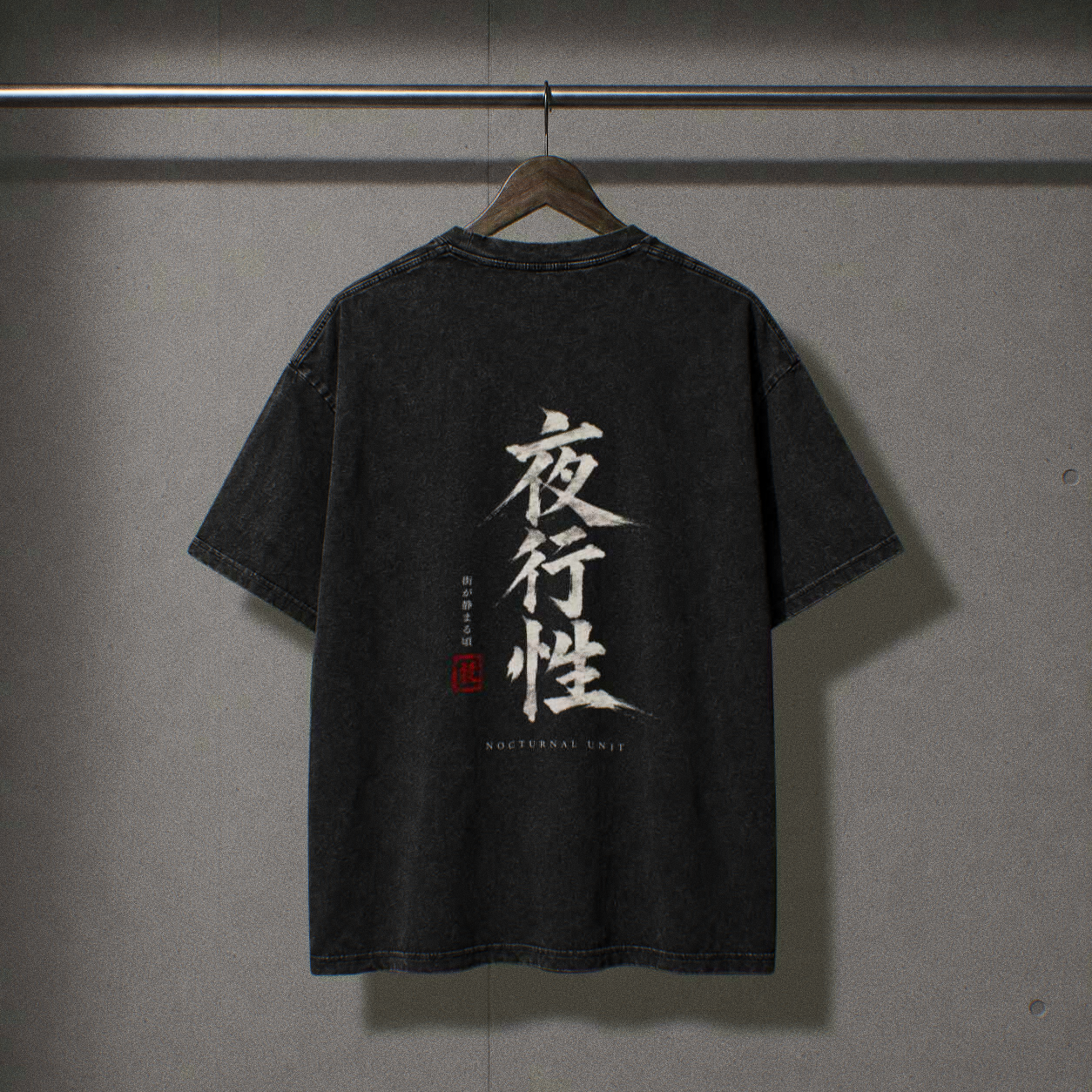 A faded black vintage washed short-sleeve t-shirt hanging on a wooden hanger against a textured gray wall. The back of the shirt features large white Japanese calligraphy characters with a red seal accent and smaller English text 'NOCTURNAL UNIT' below.