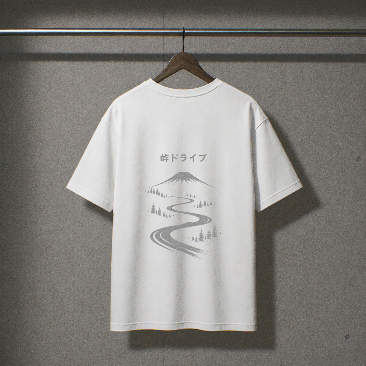 A white short-sleeve T-shirt hanging on a wooden hanger against a textured gray wall. The back of the shirt features a gray graphic print of a mountain, a winding road, and Japanese text above it.